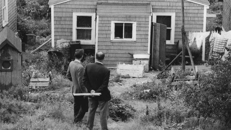 A black-and-white image of two men in suits standing in front of a wooden house with their backs to the viewer. 