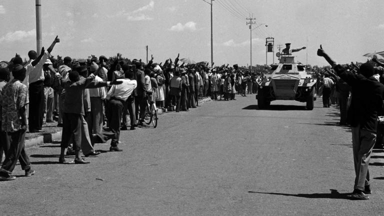 A large armoured vehicle drives down a road. Crowds on either side of the vehicle hold out their fists in a salute with their thumbs raised.