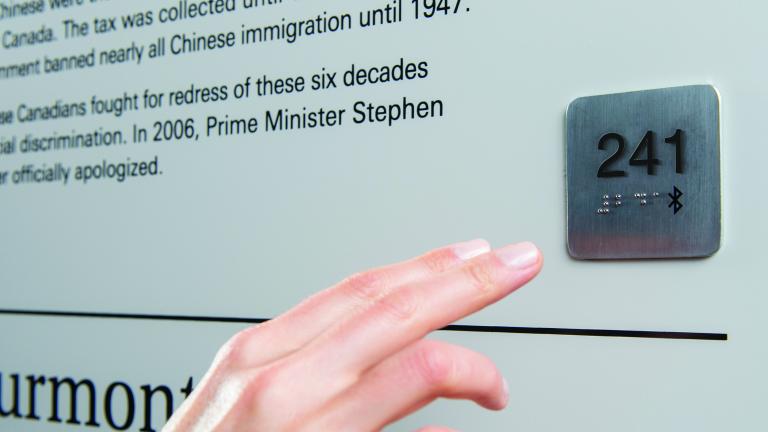 A hand reaches out to a Universal Access Point, which is a small metal square attached to a wall. The square has the number 241 imprinted on it, as well as braille writing and a Bluetooth symbol.