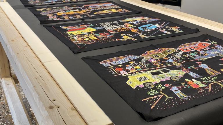 A series of colourful embroidery panels on black fabric displayed on a wooden hand-cranked conveyer belt table and on the wall of a museum.