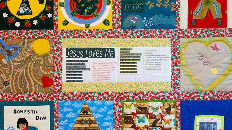 A colourful quilt with varied panels, including designs such as a bird, a heart, a teepee, a woman cleaning a floor, and lines of text under the title “Jesus Loves Me.”