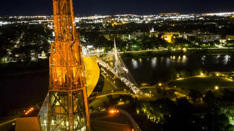 A glowing orange tower is pictured from above against a background night-time cityscape with view of a river and a bridge.