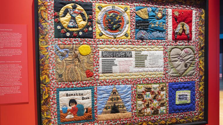 A colourful quilt, framed and hung on a red wall. The quilt includes several images including one of a girl washing a floor, a heart and Indigenous symbols.