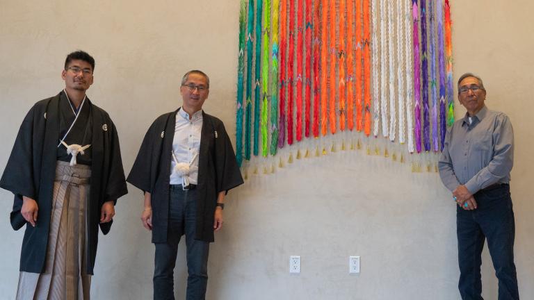Three men stand beside a colourful artwork made of one thousand folded paper cranes. Two of the men are wearing traditional Japanese clothing. The other man is wearing a jean shirt and bolo tie.