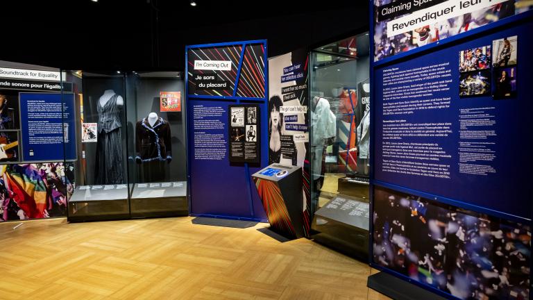 Clothing in two display cases, text and bright graphics are displayed on a standalone, curved wall with messages of 2SLGBTQI+ equality. A screen on a podium-type stand is in the centre.