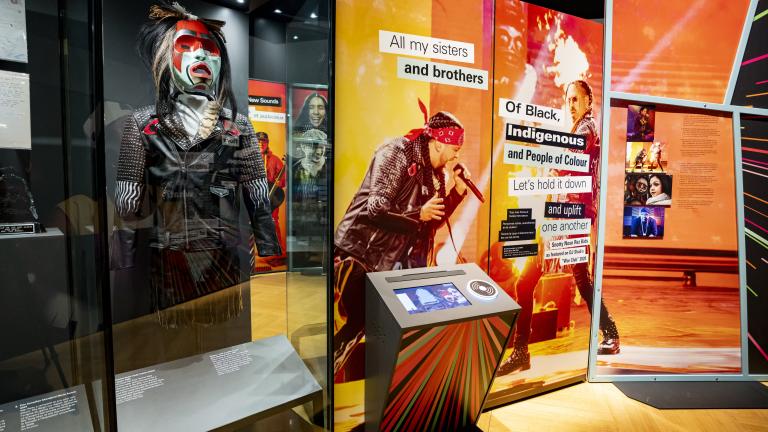 A museum exhibit highlights Indigenous musical artists. It contains a glass case displaying a mannequin wearing a mask and a jacket, as well as a screen on a podium-type structure and large graphic panel of text and images on an orange background. 