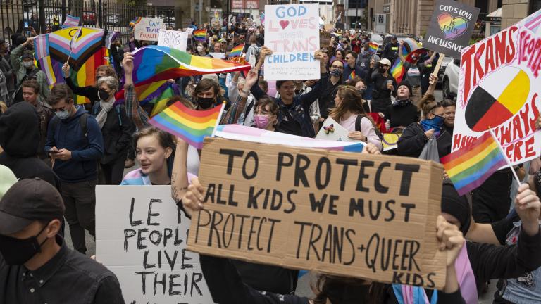 A group of people crowd into a city street holding signs and rainbow flags. The sign in the foreground reads, “To protect all kids we must protect trans and queer kids.”