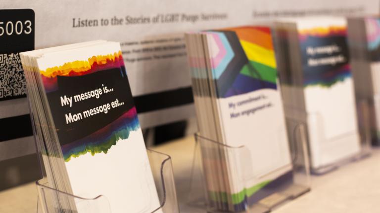 Display of cards with rainbow designs and messages, set in clear holders on a table. A poster in the background reads, “Listen to the Stories of LGBT Purge Survivors.”