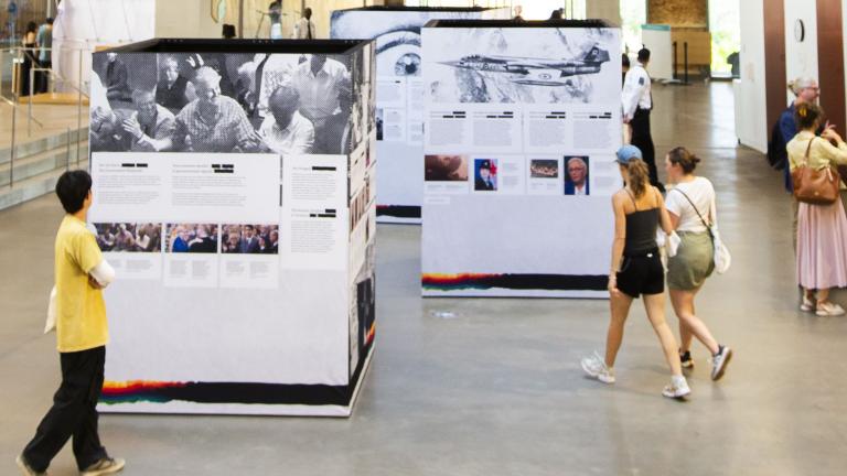 Three large cubes covered in images and text are on display in a large public room. Two groups of two adults walk near them, perusing the cubes’ content. Off to the side, four people are talking.