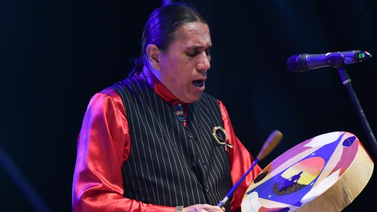 An Indigenous performer wearing a red, long-sleeve top and black, patterned vest is chanting and drumming.