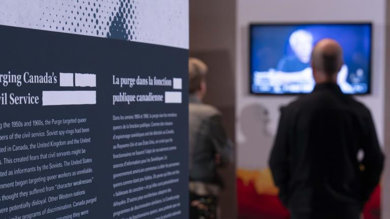 An exhibit panel with the text “Purging Canada’s Civil Service” is on the left. Two people in the background on the right watch a video. 