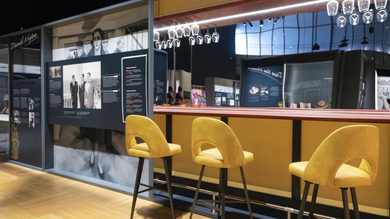 A replica of a small bar featuring high yellow stools, a mirrored wall behind the bar, and wine glasses hanging from overhead racks is next to an exhibition panel with a large image of people and text.