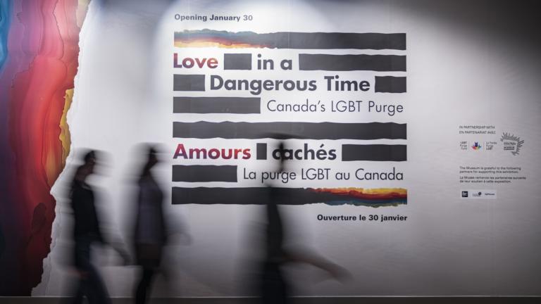A small group of people walk by a large sign on a wall, made up of thick black horizontal lines with the English and French words “Love in a Dangerous Time: Canada’s LGBT Purge” and “Amours cachés : La purge LGBT au Canada” distributed within the lines. The top and bottom lines are a mix of black and rainbow colours. Rainbow colours cover the left edge of the wall.