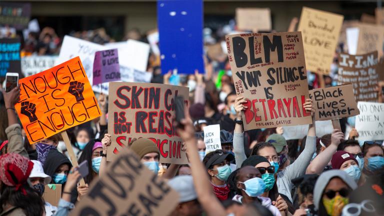 A large group of people holding signs with slogans such as “Black Lives Matter,” “Enough is Enough” and “Your Silence is Betrayal.” Partially obscured.
