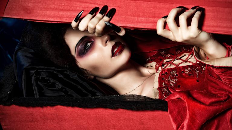 A vampire dressed in a velvety red dress peeks through a red wooden box with her long, black nails. Partially obscured.
