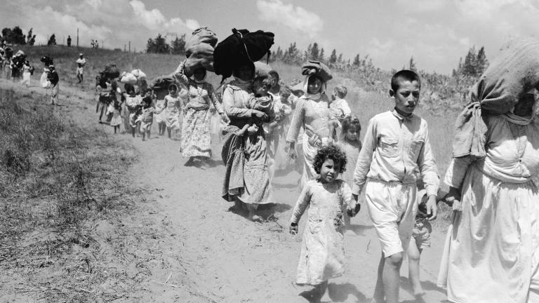 Women and children carrying bundles on their heads walk along a dirt road. Partially obscured.