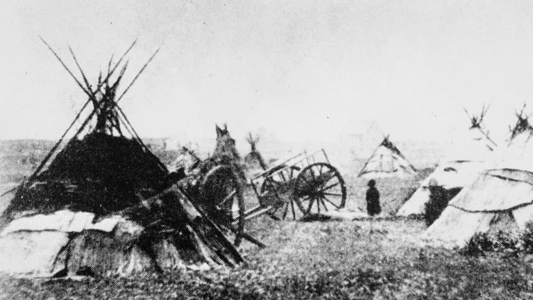A grainy black and white photo of several tipis and a wooden cart in a field. Partially obscured.