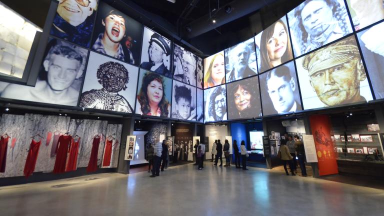 The museum's Canadian Journeys gallery, seen from the perspective of a visitor standing in the middle of it, surrounded by niches that share stories of Canadian experiences of human rights. On the walls above the niches, large images of faces of Canadian human rights defenders are seen arranged in a grid pattern. Partially obscured.