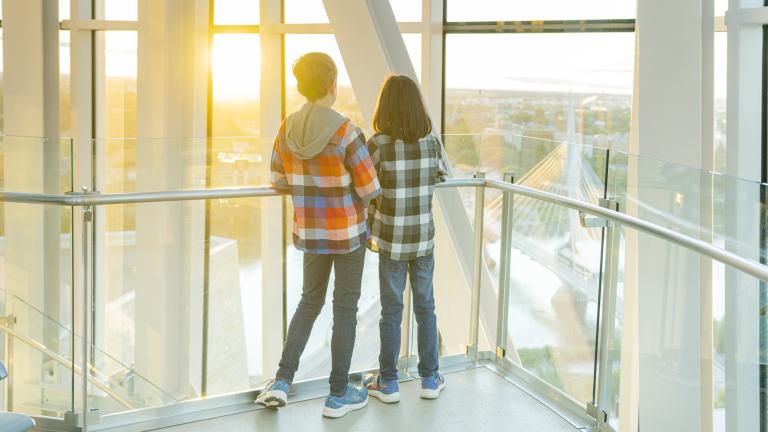 Two young children stand in a tower with their backs toward the camera as they stare out at a horizon overlooking a cityscape. Partially obscured.