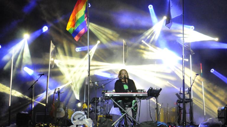 A performer is sitting behind a keyboard on stage with beams of blue and white lights and a rainbow flag flying on a pole. Partially obscured.