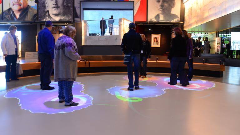 A group of people are standing within part of a white circle. Blue and purple lights are projected onto the floor surrounding each person. Partially obscured.