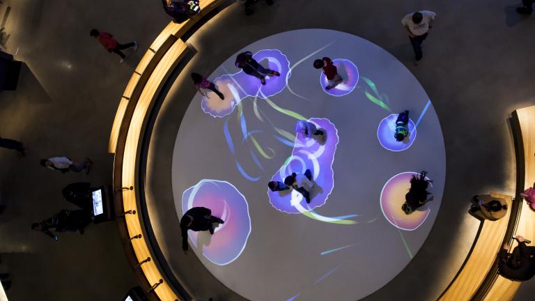An overhead view of people walking on a white circle on the floor. Each person is surrounded by a bubble of projected, colourful light. Partially obscured.