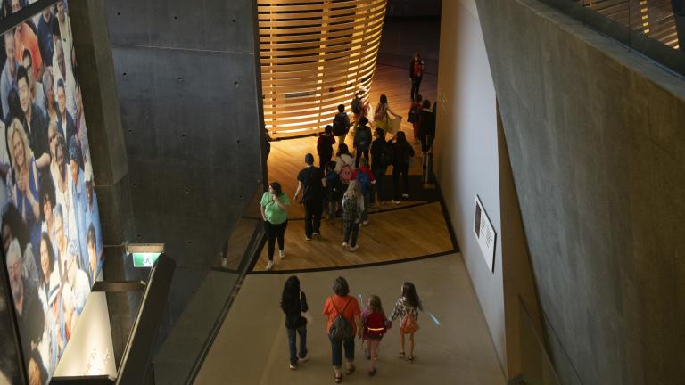Students walking through Museum galleries. On the left wall is an image of a crowd of people. The walls are made of cement. Partially obscured.