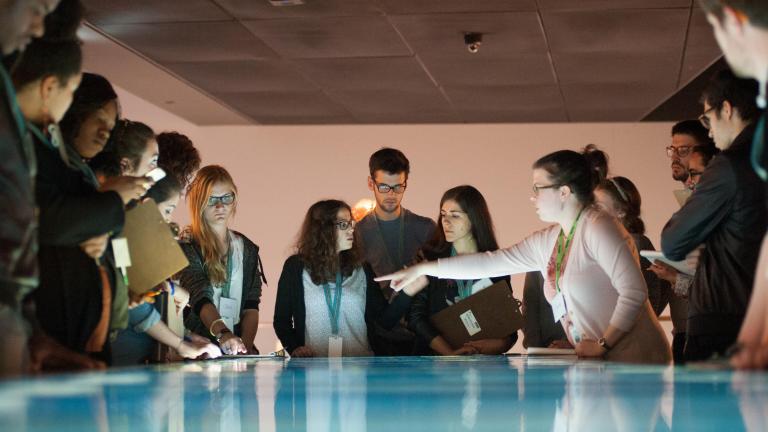 A group of young people stand around an interactive table emitting blue and white light. Partially obscured.