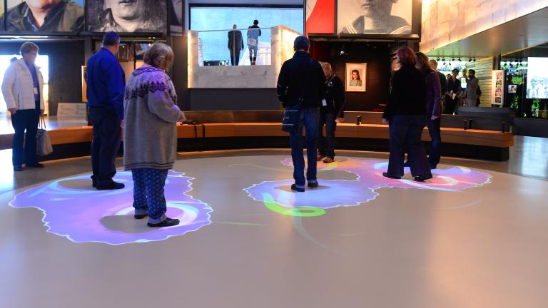 A group of people are standing within part of a white circle. Blue and purple lights are projected onto the floor surrounding each person. Partially obscured.