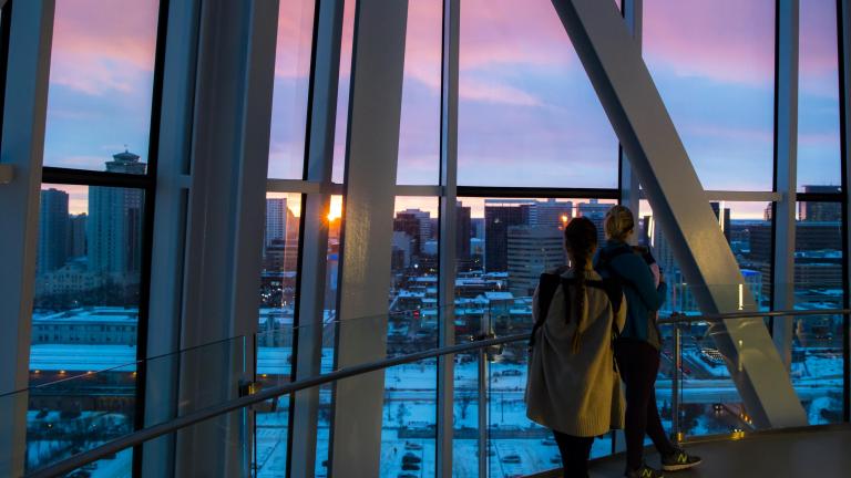 Two individuals stand looking through the floor-to-ceiling glass windows at the Winnipeg skyline. The sun is setting. Partially obscured.
