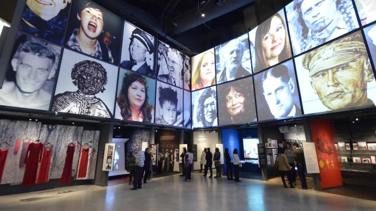 People explore a Museum gallery with two rows of square portraits above a series of alcoves, containing photographs, videos, and text. The alcove on the left contains suspended red dresses. Partially obscured.
