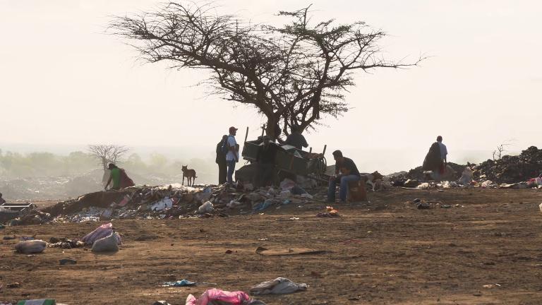 Several people and one dog sitting and standing in a landfill. A tree sits in the centre of the image and garbage is strewn on the ground. Partially obscured.