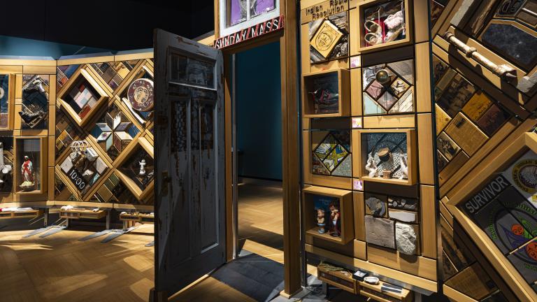 A large, free-standing art installation contains hundreds of objects. It is framed in cedar and there is an open door in the middle that people can walk through. Partially obscured.