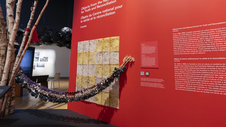 A museum exhibit featuring many traditional Indigenous medicine bags hanging from a baby swing. One end of the swing is attached to birch tree trunks and the other to a red wall that includes text panels. Partially obscured.