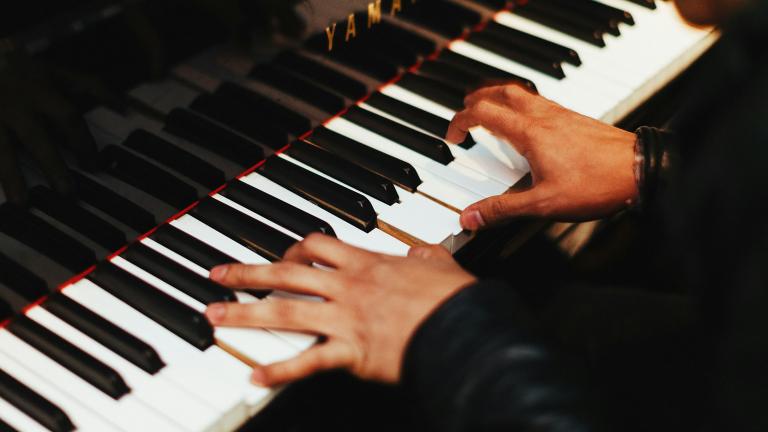 Two hands play the keys of a gleaming black Yamaha piano. The pianist, barely shown, wears a long-sleeved black top. Partially obscured.