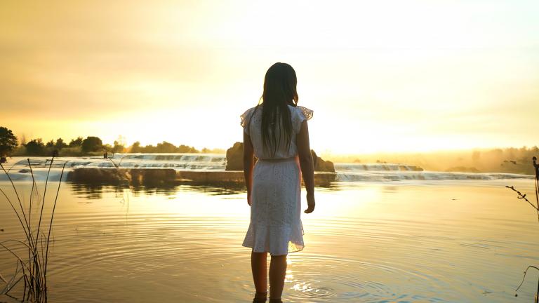 A girl with long dark hair wearing a knee-length white dress faces away, standing ankle deep in a calm lake, with the setting sun casting a warm yellow glow across the water. Partially obscured.