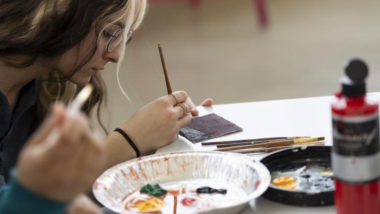 A young woman sitting at a table paints a design on a tile while a tin tray with various paints sits in front of her. An open bottle of red paint sits on the table. Partially obscured.