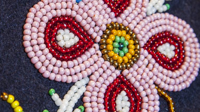 Close-up of a beaded floral design on dark fabric, featuring a pink four petal flower with red and white details, a yellow and green beaded center, and surrounding beadwork resembling leaves and stems. Partially obscured.