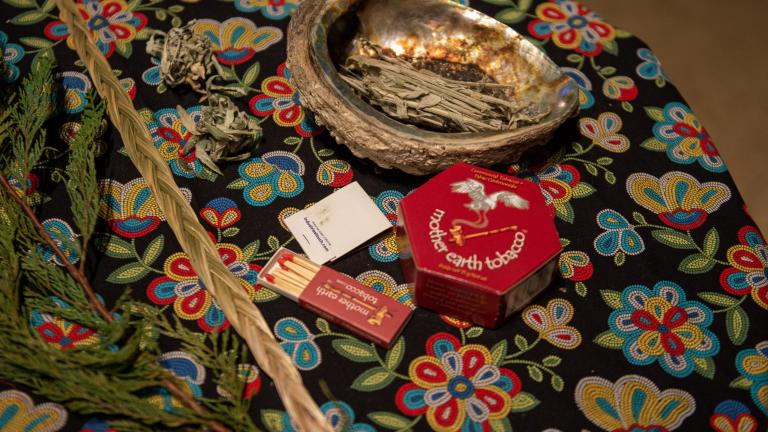 Smudging items arranged on a round table covered with a floral cloth, including an abalone shell holding dried sage, a feather, braided sweetgrass, cedar branches, and a small box of matches. Partially obscured.