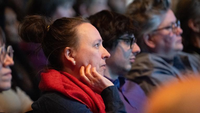 Audience members sit in a dimly lit venue watching a presentation, their faces illuminated by stage light. In the foreground, a person with their hair in a bun and wearing a bright red scarf rests their chin on their hand, looking attentively toward the stage. Other attendees sit beside them in soft focus, creating a quiet, contemplative atmosphere as the crowd listens. Partially obscured.