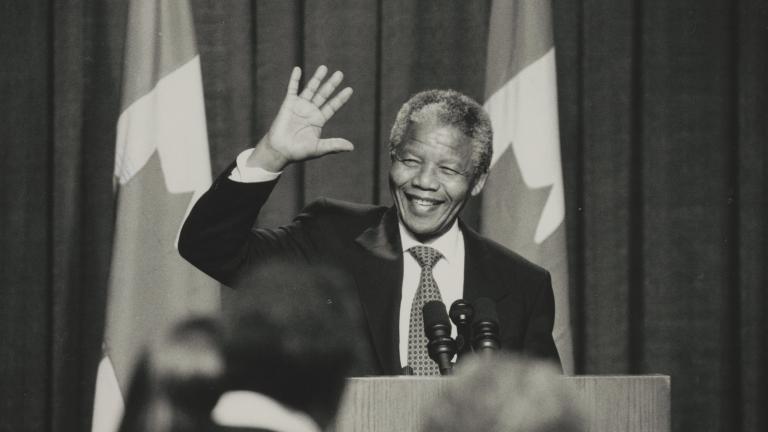 A black and white photograph of Nelson Mandela standing in front of a podium. He is smiling and waving. Partially obscured.