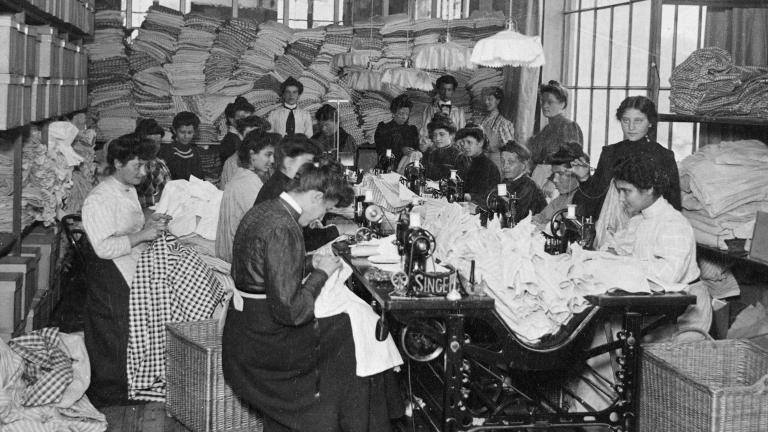 A large group of women sitting and sewing. Others are standing behind them in a room crowded with textiles. Partially obscured.