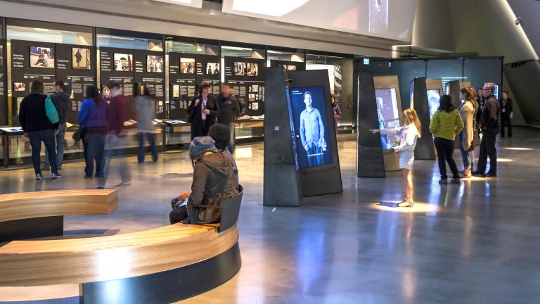 People explore a gallery containing vertical interactive videos in the centre of the room; and displays containing text and images that line the back wall.