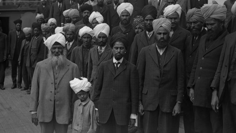 A black and white image of a large group of Sikh men and one young boy standing on the deck of a ship. They are all facing the camera and are not smiling.