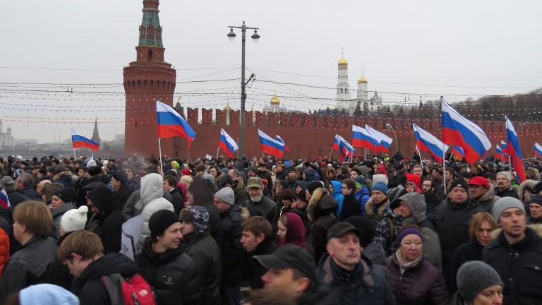A crowd of people marches holding the red, white and blue striped flags of Russia. Behind is a red concrete wall that ends in a spire.