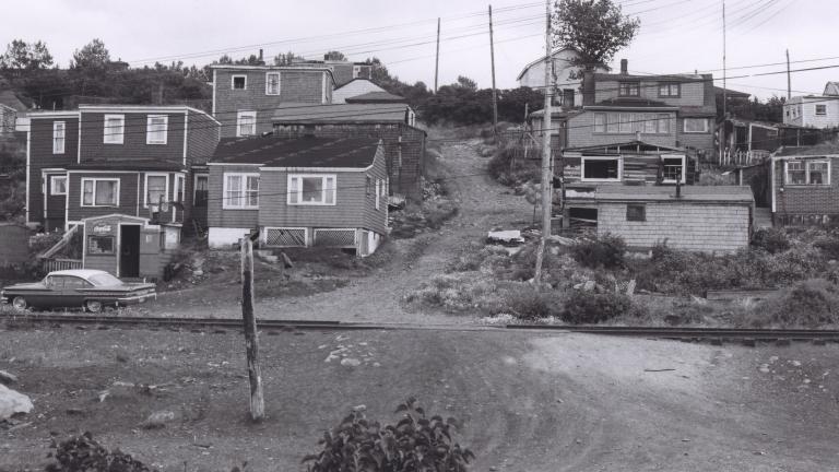 A black and white image of about a dozen houses on a hill. In the middle of the image, a dirt road travels up the hill between the houses.