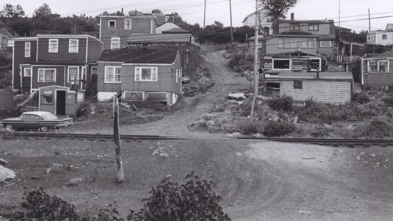A black and white image of about a dozen houses on a hill. In the middle of the image, a dirt road travels up the hill between the houses. Partially obscured.