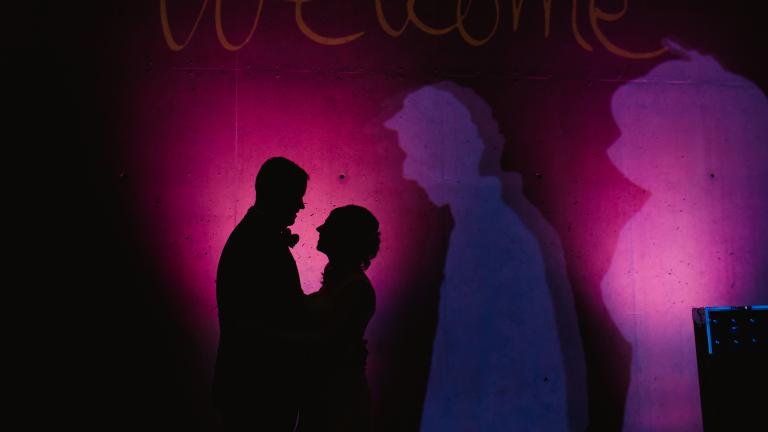 A man and woman in silhouette face each other in front of a dark wall illuminated by purple light. Partially obscured.