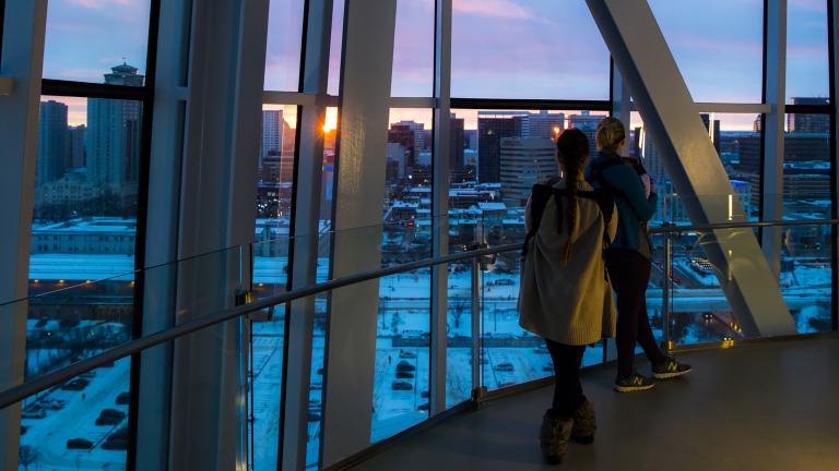 Two people gaze out a tall glass window at a sunset over a cityscape. Partially obscured.