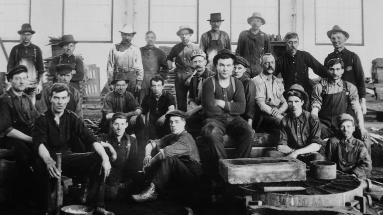 A group of men in working clothes sitting and standing in a factory.